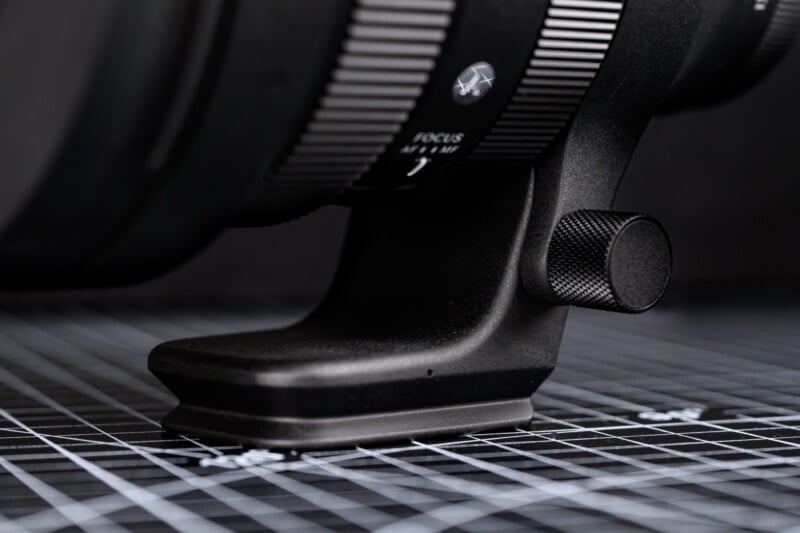 Close-up of a camera lens tripod mount resting on a black and white grid-patterned surface, showing detailed texture and focus adjustment markings.