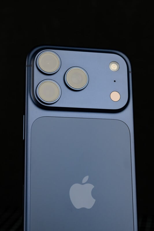 A close-up of the back of a blue iPhone showing three camera lenses, a flash, and the Apple logo against a dark background.