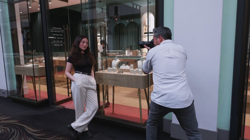 A photographer takes a photo of a woman posing against a jewelry store's window display. The woman is wearing a black top and white pants with hands in pockets, while the photographer is crouched and aiming his camera at her.