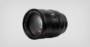 A black camera lens with detailed markings and switches is displayed on a light gray background, showing its glass elements and various control dials.