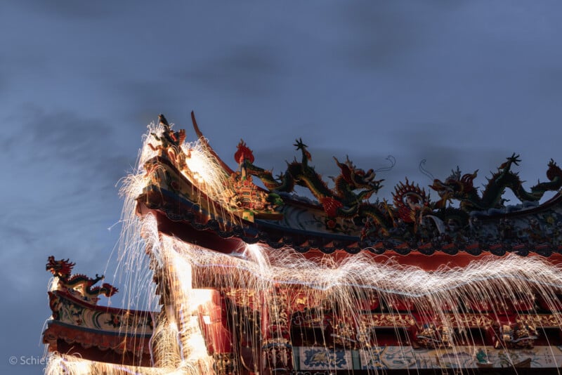 Fireworks shoot sparks over a traditional Asian temple roof adorned with ornate dragon sculptures, set against a dusky sky. The long exposure creates trails of light that sweep across the rooftop.