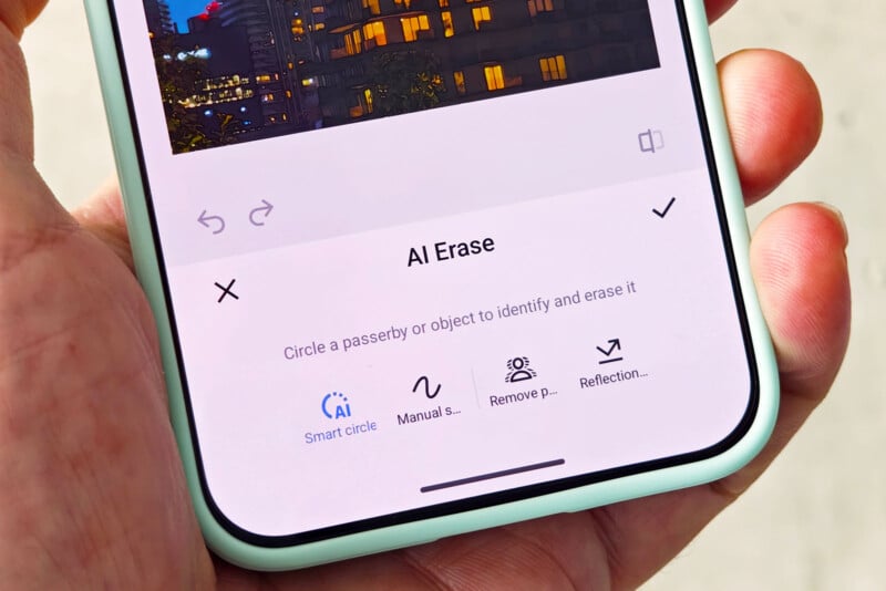 A hand holding a smartphone displaying a photo editing app with the “AI Erase” tool open, offering options to erase objects, including Smart circle, Manual select, Remove person, and Reflection erase.