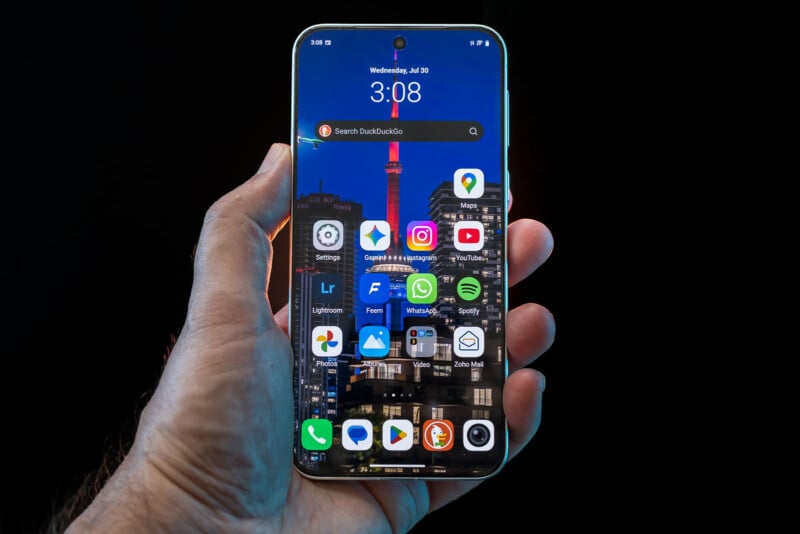 A hand holding a smartphone displaying various app icons on the home screen, with a cityscape wallpaper in the background. The time on the phone is 3:08.