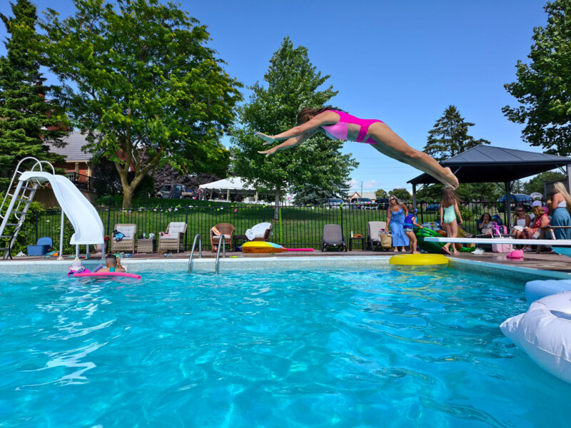 A woman in a pink swimsuit dives off a diving board into a clear blue swimming pool as people, including children, watch and play nearby on a sunny day. Trees and lawn chairs are visible in the background.