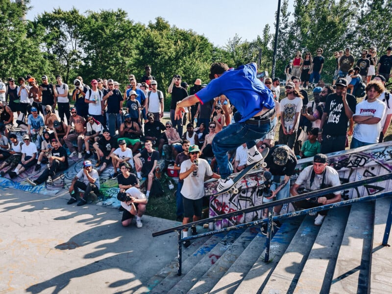 A skateboarder performs a trick over a handrail at a crowded outdoor skatepark, surrounded by spectators watching and taking photos on a sunny day.