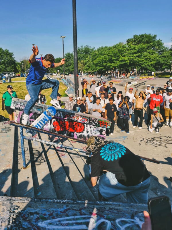 A skateboarder performs a trick on a rail covered in graffiti while a crowd watches and a photographer crouches nearby at a sunny outdoor skate park.