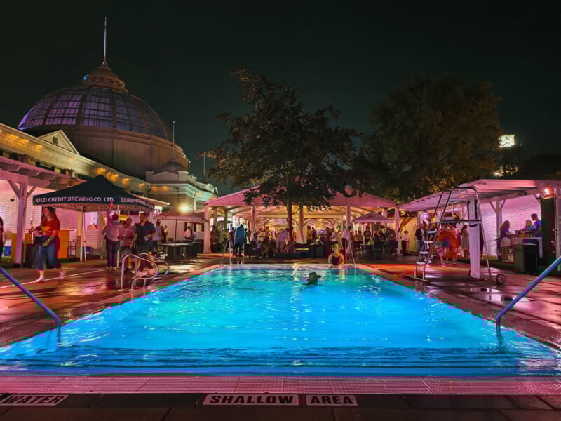 A brightly lit outdoor pool at night, surrounded by people socializing under canopies. Trees and a domed building are visible in the background, with colorful lights reflecting on wet surfaces.