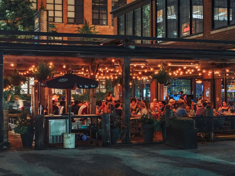 A lively outdoor restaurant patio at night, filled with people dining and talking under warm string lights, surrounded by plants and brick buildings.