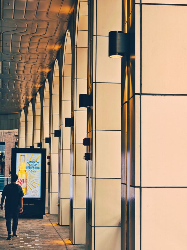 A person walks along a covered walkway lined with tall, cream-colored arches and modern wall lights. A bright advertisement is visible ahead, and sunlight highlights the geometric patterns on the ceiling and walls.