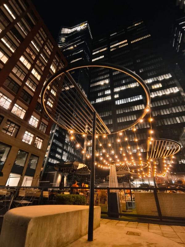 Outdoor dining area at night with string lights hanging in circular patterns, surrounded by tall modern buildings with illuminated windows. The atmosphere appears lively and urban.