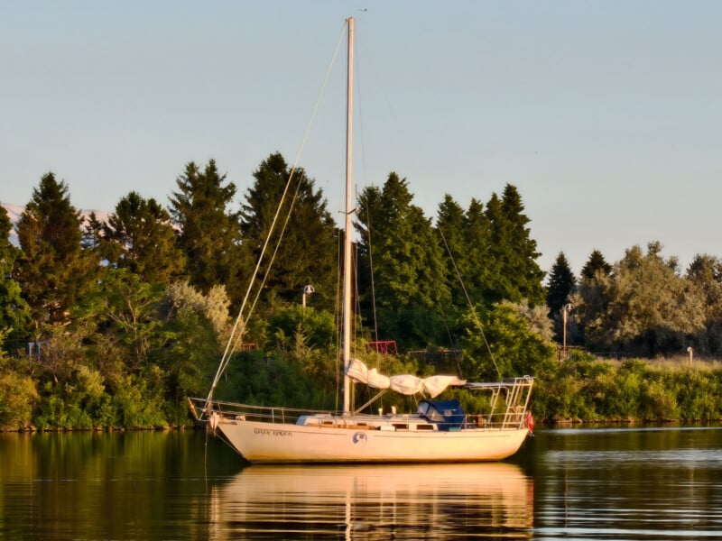 A cream-colored sailboat floats on calm water near a lush, tree-lined shore under warm sunlight, with its reflection visible on the water’s surface.