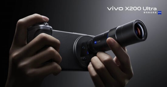 Close-up of hands holding a Vivo X200 Ultra smartphone with an attached external lens, highlighting advanced photography features. The phone is black and the lens is prominently visible.