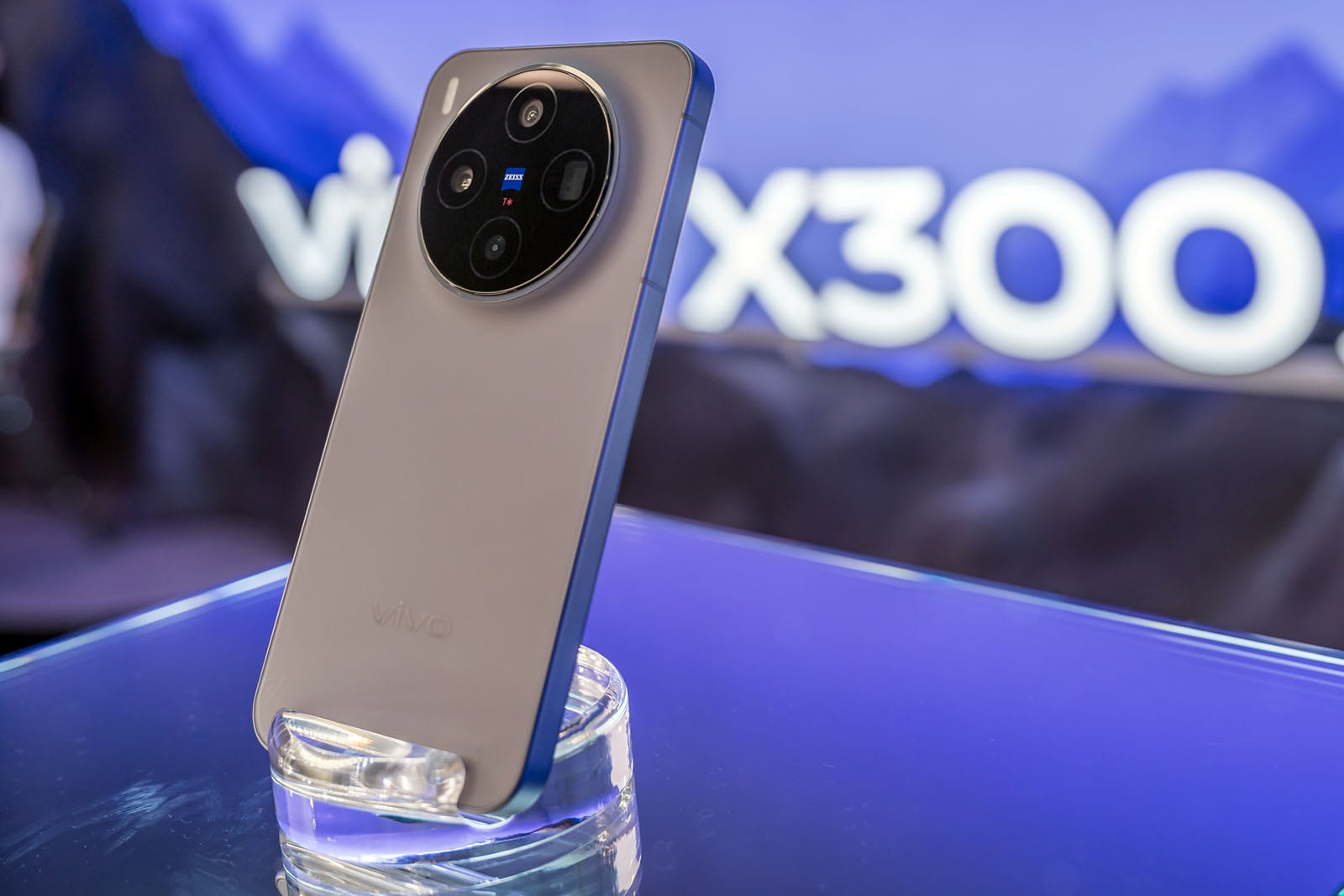 A silver Vivo smartphone with a large circular rear camera module is displayed on a clear stand, with blurred "Vivo X300" branding in the blue-lit background.