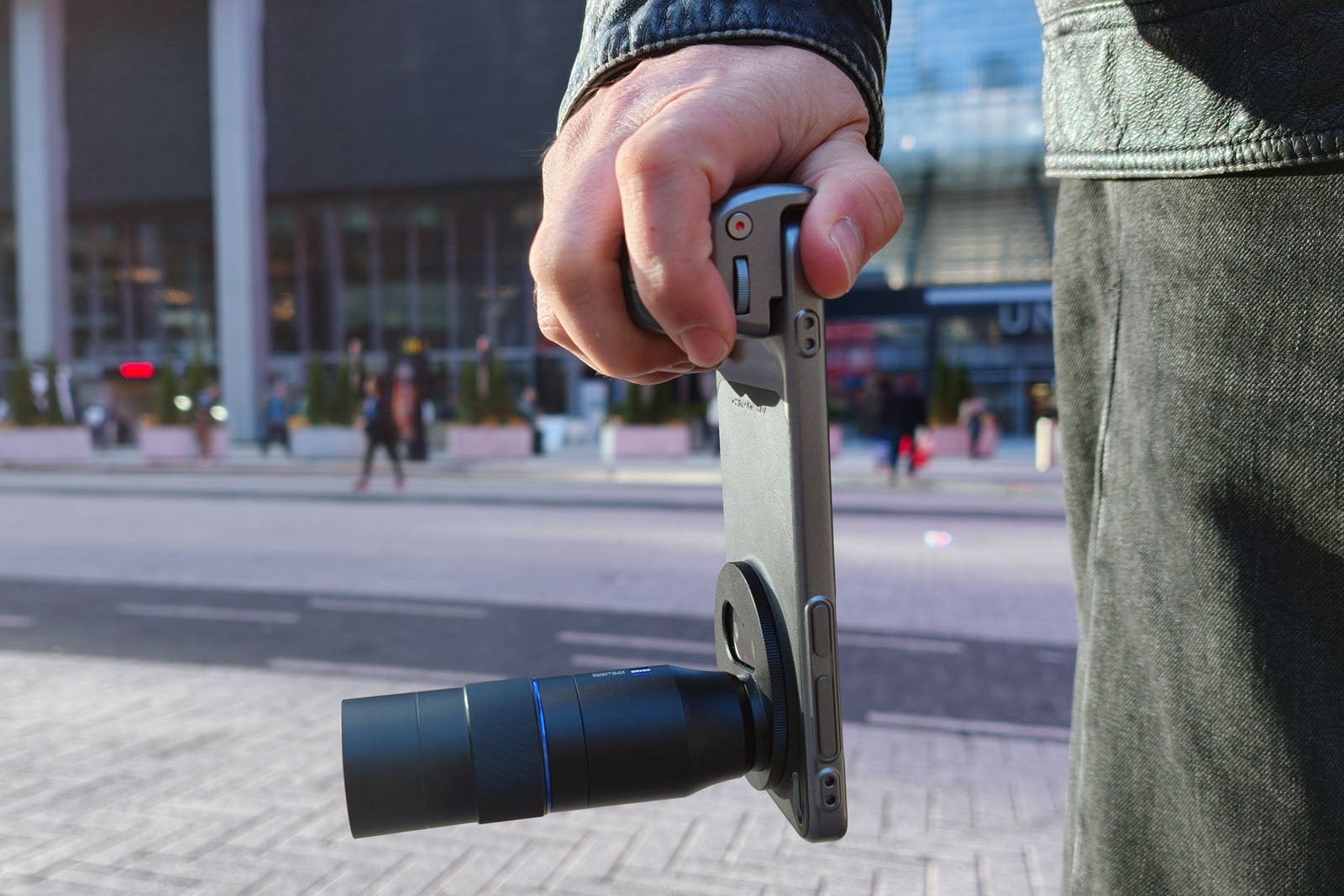 A person holding a smartphone with an attached external camera lens, pointing it toward the street while standing outdoors in an urban area.