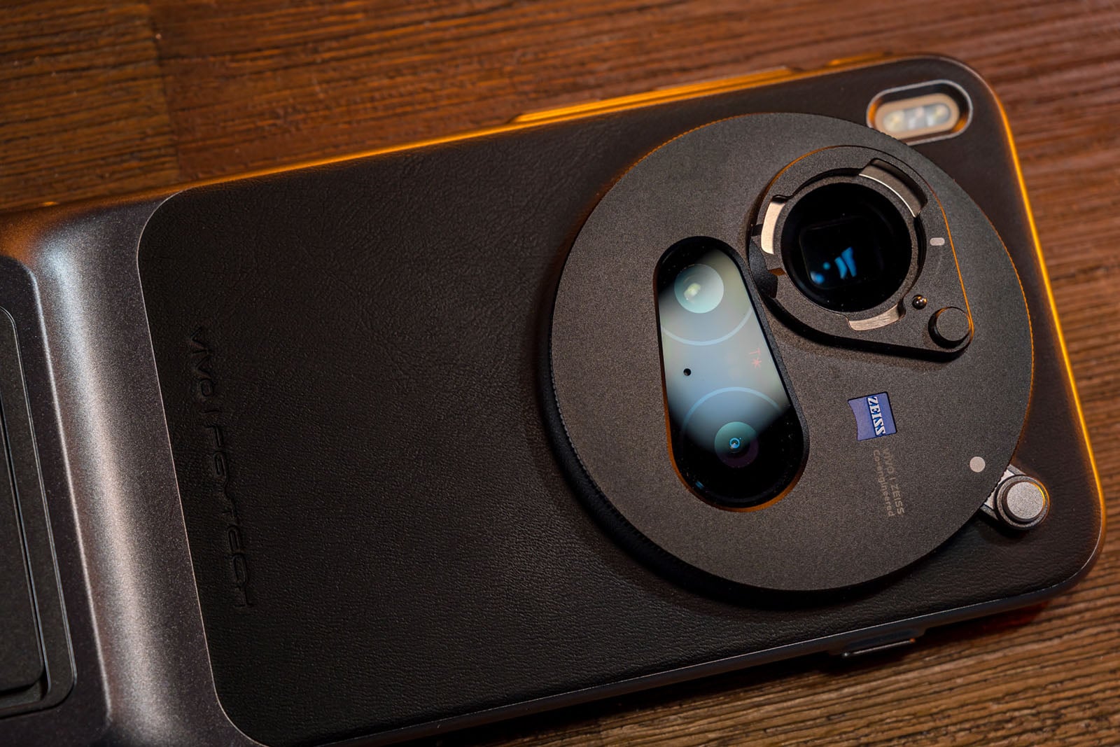 Close-up of a smartphone with a large circular camera module featuring Zeiss branding, dual camera lenses, and a textured black case, placed on a wooden surface.