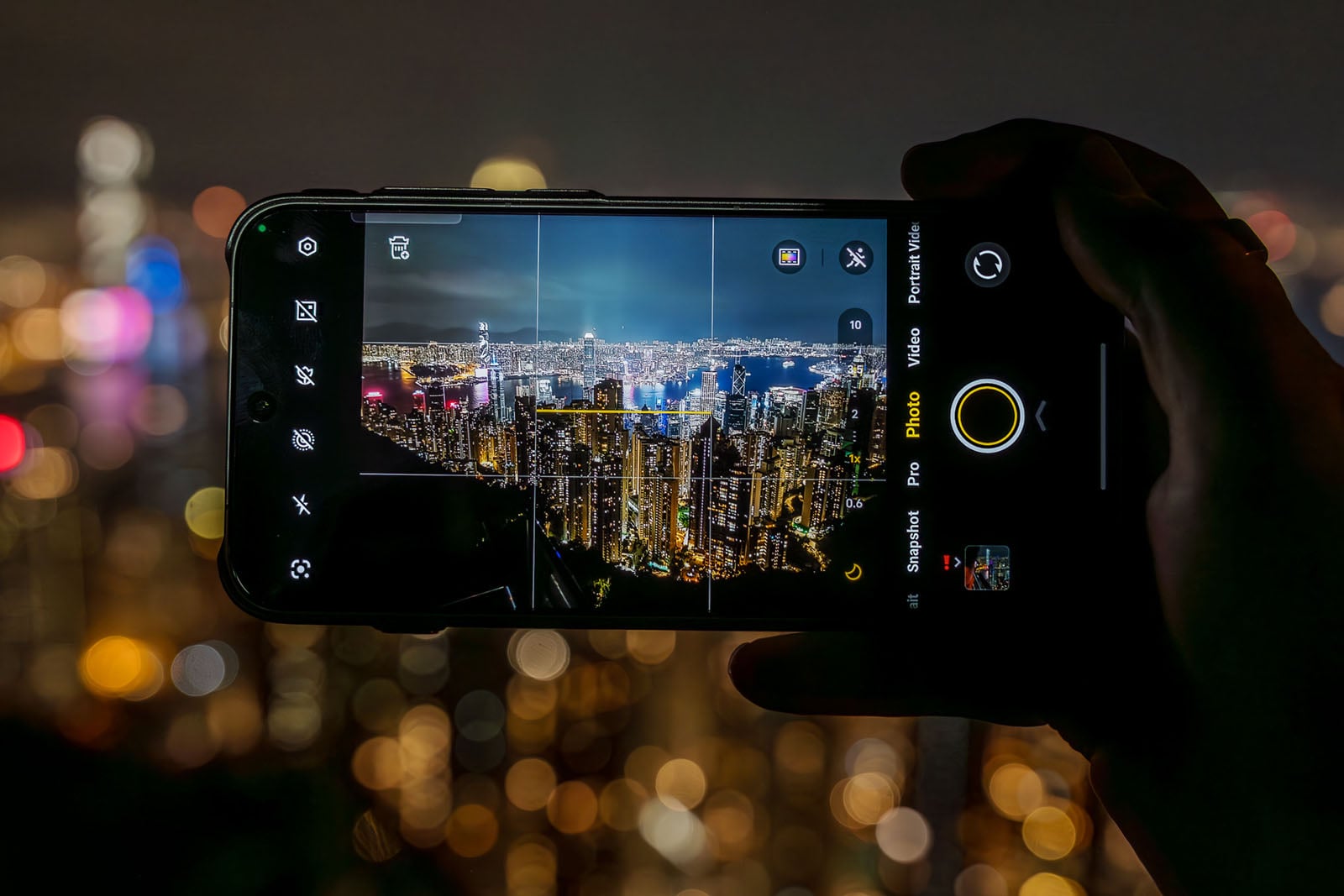 A hand holds a smartphone, capturing a photo of a city skyline at night. The phone screen displays the brightly lit cityscape in focus, while the background appears blurred with colorful lights.
