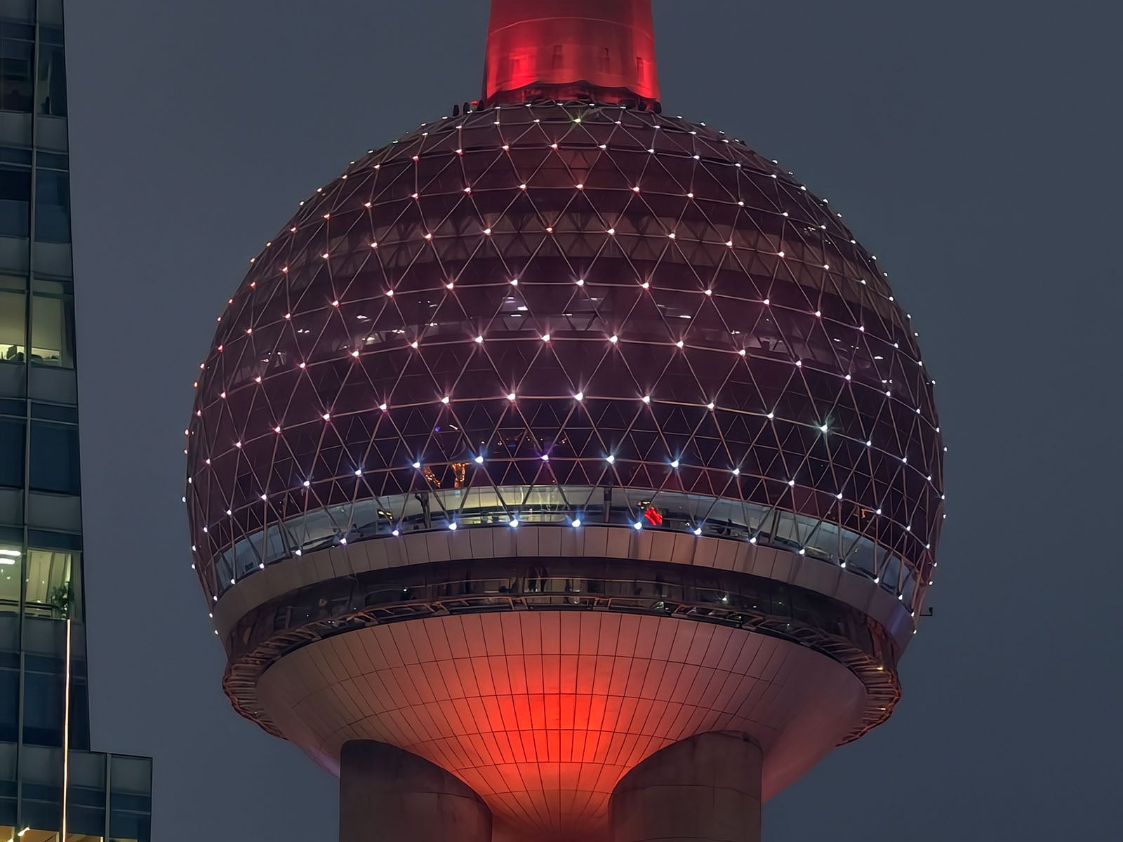 A close-up view of a large spherical tower with a lattice pattern illuminated by red and white lights at night, partially framed by a modern glass building on the left.