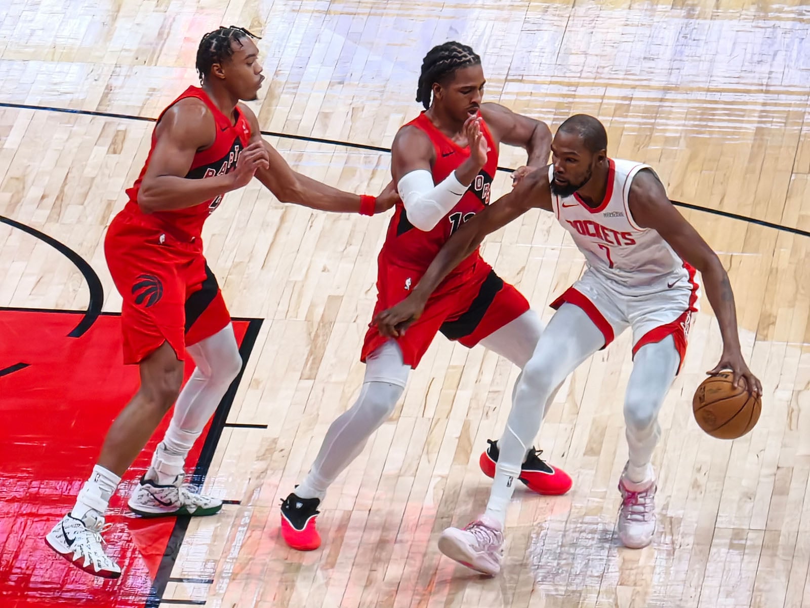 A Houston Rockets basketball player dribbles the ball while being closely guarded by two Toronto Raptors players on a basketball court during a game.