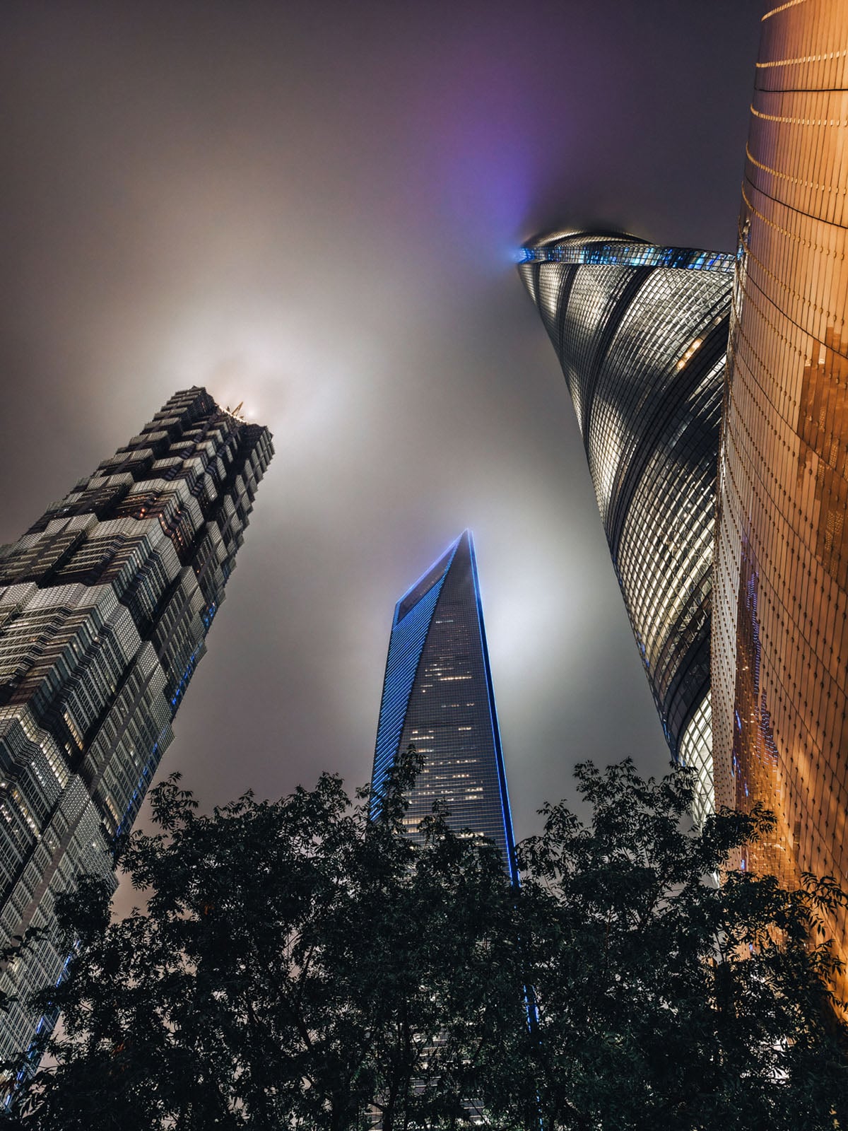 Three modern skyscrapers rise into a foggy night sky, their tops illuminated by lights and partially obscured by mist, with tree branches visible at the bottom of the image.