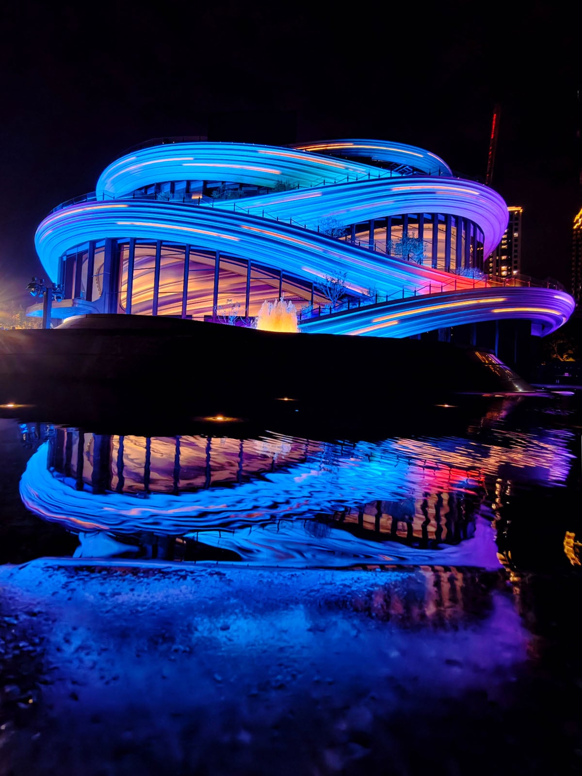 A modern building with swirling, ribbon-like architecture is illuminated by vibrant blue, purple, and pink lights at night, with colorful reflections shimmering in the water below.