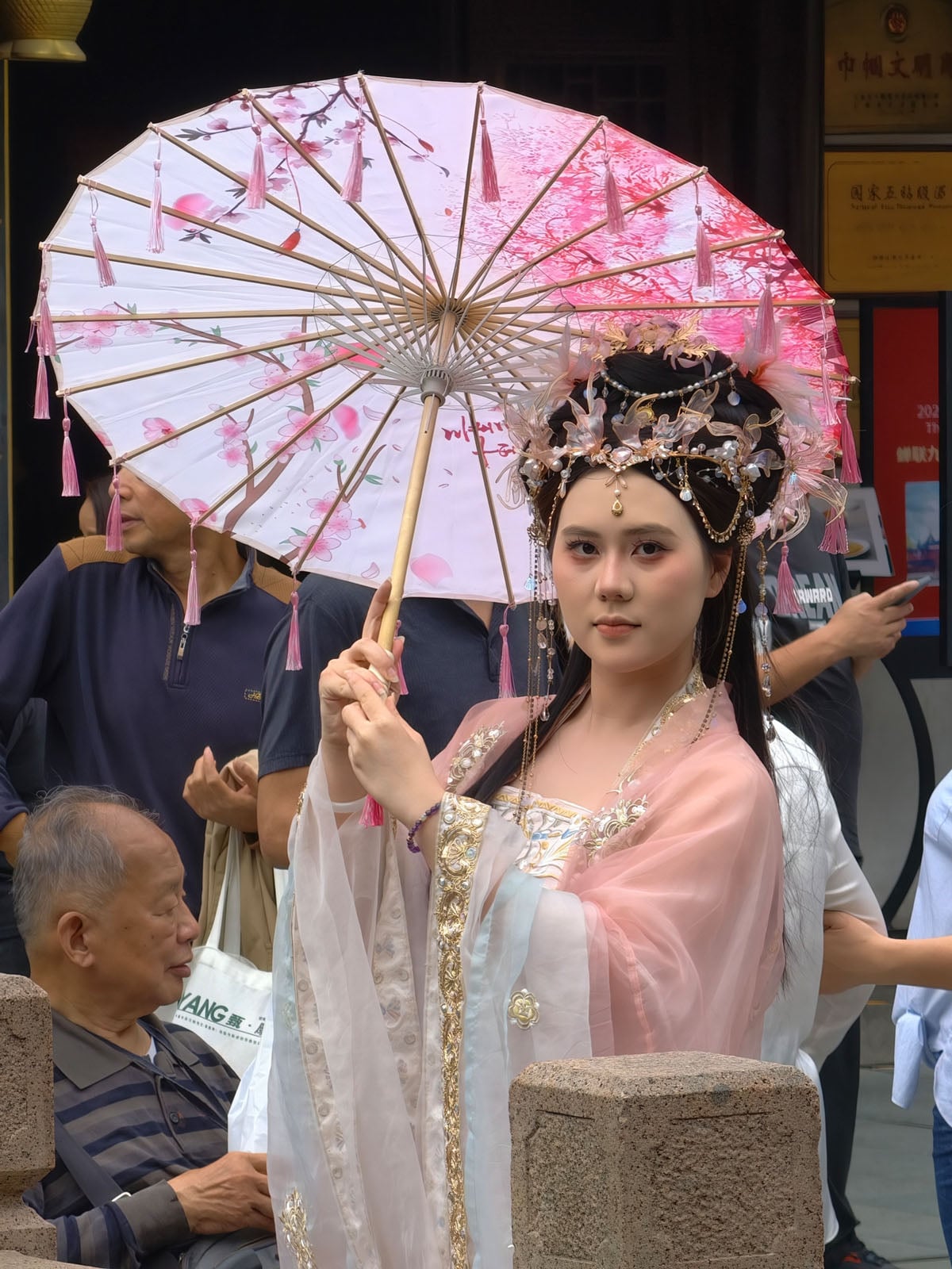 A woman in traditional Chinese dress and ornate headpiece holds a decorative pink parasol with tassels. She stands among a crowd, looking at the camera. The scene appears to be at a public event or festival.
