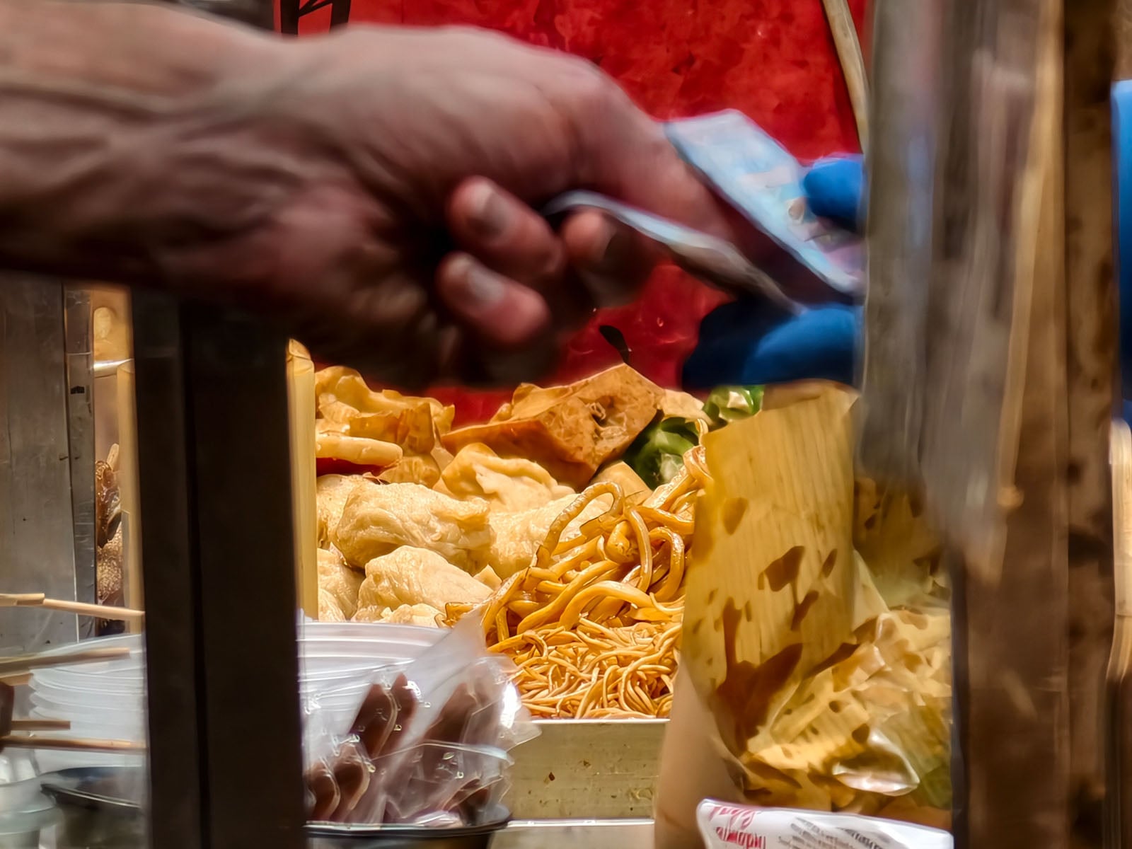 A close-up of hands exchanging cash at a food stall with fried noodles and various fried snacks in the background. One hand holds money, while the other, wearing a blue glove, receives it near a takeout bag.