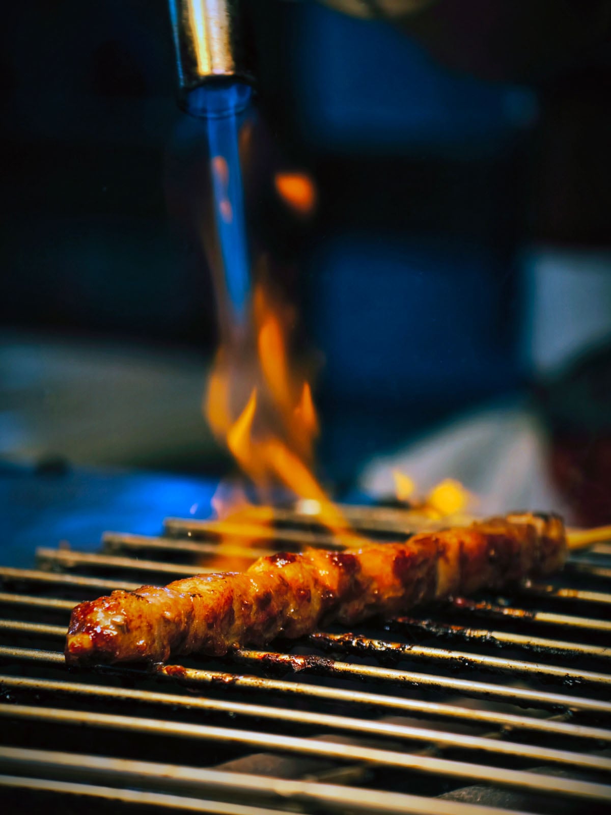 A skewer of meat is being grilled over an open flame, with a torch adding direct heat to the meat. The grill bars are visible, and the scene is dramatic with bright orange flames and a dark background.
