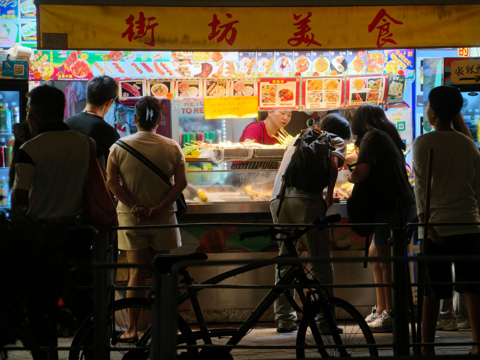 A group of people stand in front of a brightly lit street food stall at night, examining the menu and ordering food, with a bicycle parked in the foreground.