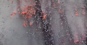 Bright orange lava splatters and flows down a dark, ash-covered surface, creating a vivid contrast between the glowing molten rock and the charred surroundings.