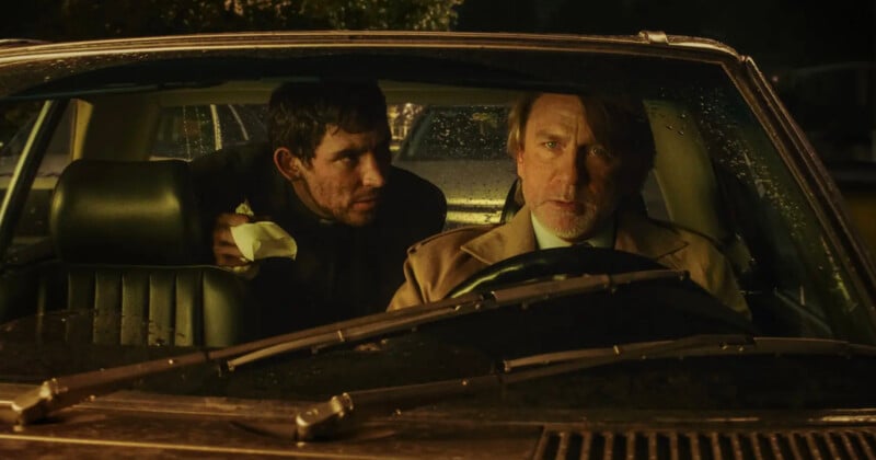 Two men sit in a car at night during rain; the man in the driver's seat looks forward seriously, while the man in the passenger seat leans forward, holding a napkin and staring at the driver.