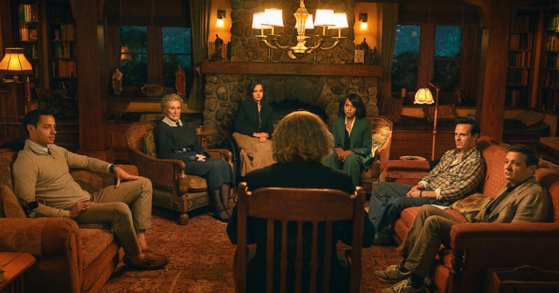 Seven people sit in a warmly lit living room arranged in a circle around a stone fireplace, listening to someone with their back to the camera. The atmosphere is serious and contemplative.