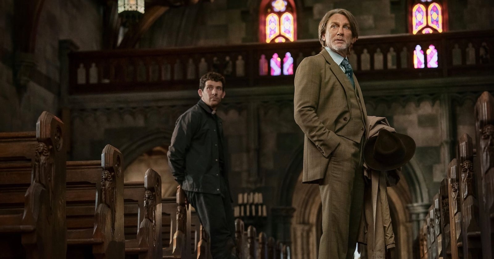 Two men stand in the aisle of an old, dimly lit church with wooden pews and stained glass windows; one wears a brown suit holding a hat, the other wears dark clothes and looks towards him.