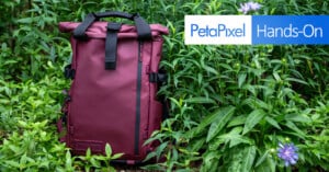 A maroon backpack stands upright among lush green plants, with purple flowers nearby. An overlay in the top right reads "PetaPixel Hands-On" in blue and white text.