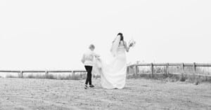 A bride in a white dress holding a bouquet walks outdoors on a sandy path, while a young boy helps carry her train. A wooden fence and grass are visible in the background. The scene is in black and white.
