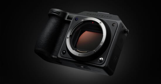 A close-up view of a Hasselblad mirrorless camera body without a lens, shown at an angle against a dark background. The camera's textured grip and lens mount are clearly visible.