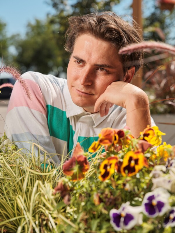 A young man with brown hair in a colorful striped shirt rests his chin on his hand and gazes thoughtfully at vibrant pansies and grasses in a sunny outdoor garden setting.