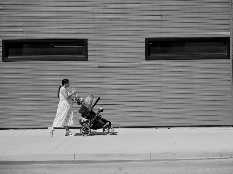 A woman in a long dress pushes a baby stroller along a sidewalk beside a modern building with horizontal siding and two rectangular windows. The image is in black and white.