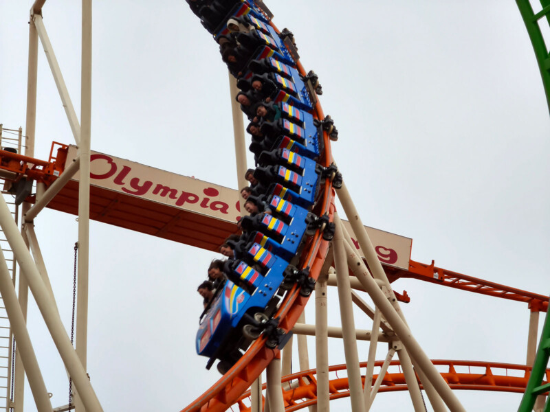 A blue roller coaster car filled with people speeds through a vertical loop on a roller coaster track, with a sign reading "Olympia" visible in the background against a cloudy sky.
