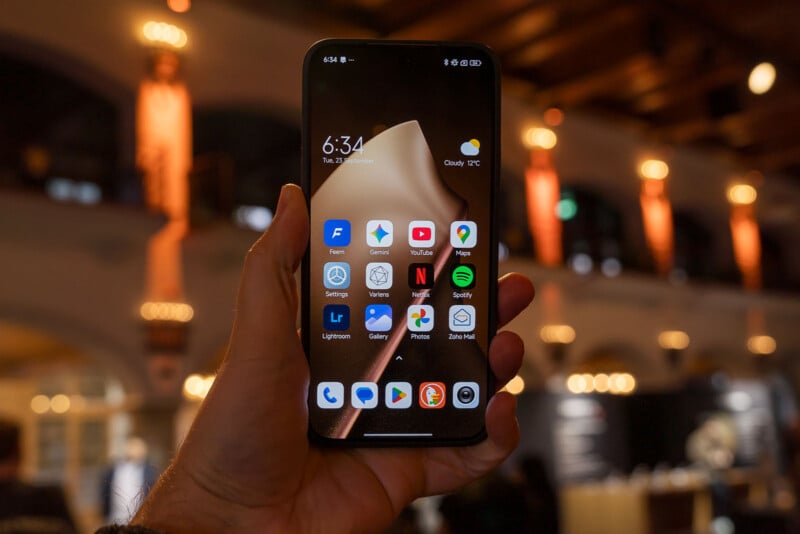 A hand holding a smartphone displaying various app icons, such as YouTube, Maps, Gmail, and Netflix, against a warmly lit indoor background with blurred lights and architectural elements.