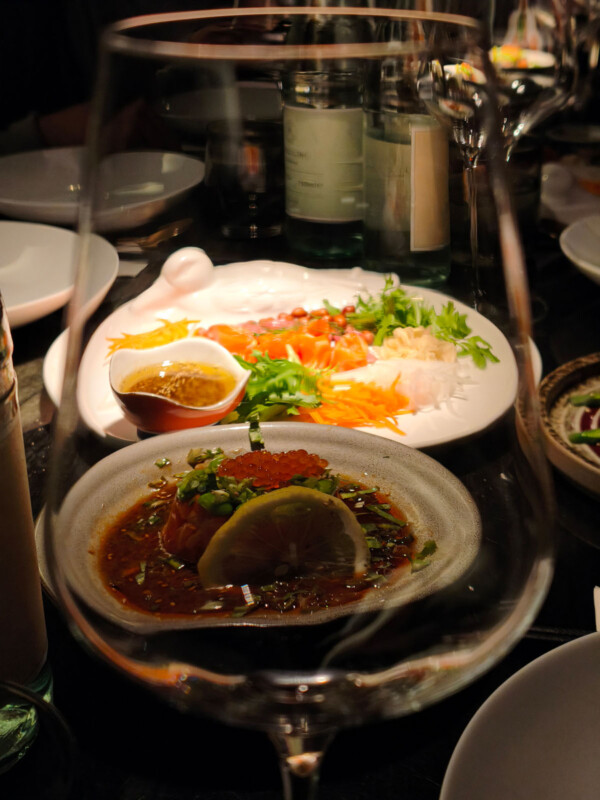 A close-up view of a dining table seen through a wine glass, featuring a plate of sashimi with greens and sauce, and another dish garnished with lemon and herbs, with bottles and plates in the background.
