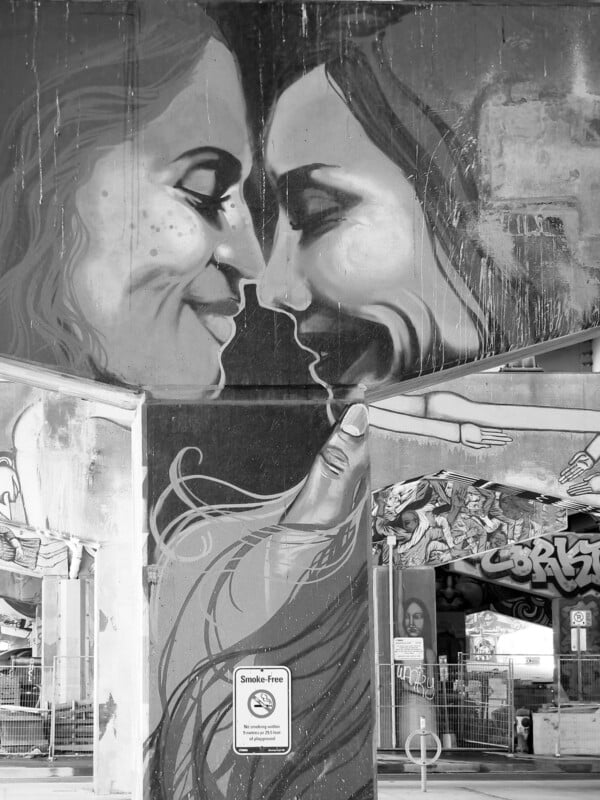 A large mural under a bridge shows two people smiling and touching foreheads, with a hand below them. Surrounding walls are covered in graffiti. A "Smoke-Free" sign is posted on a column in the foreground. The image is in black and white.