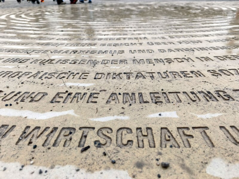 Close-up of German text engraved in stone on a wet, circular monument. The surface shows raindrops and reflections, with blurred people visible in the background.