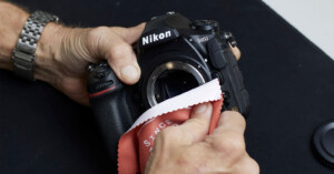 A person uses a red microfiber cloth to clean the sensor area of a Nikon DSLR camera, holding the camera in one hand and wiping with the other. The camera lens has been removed.
