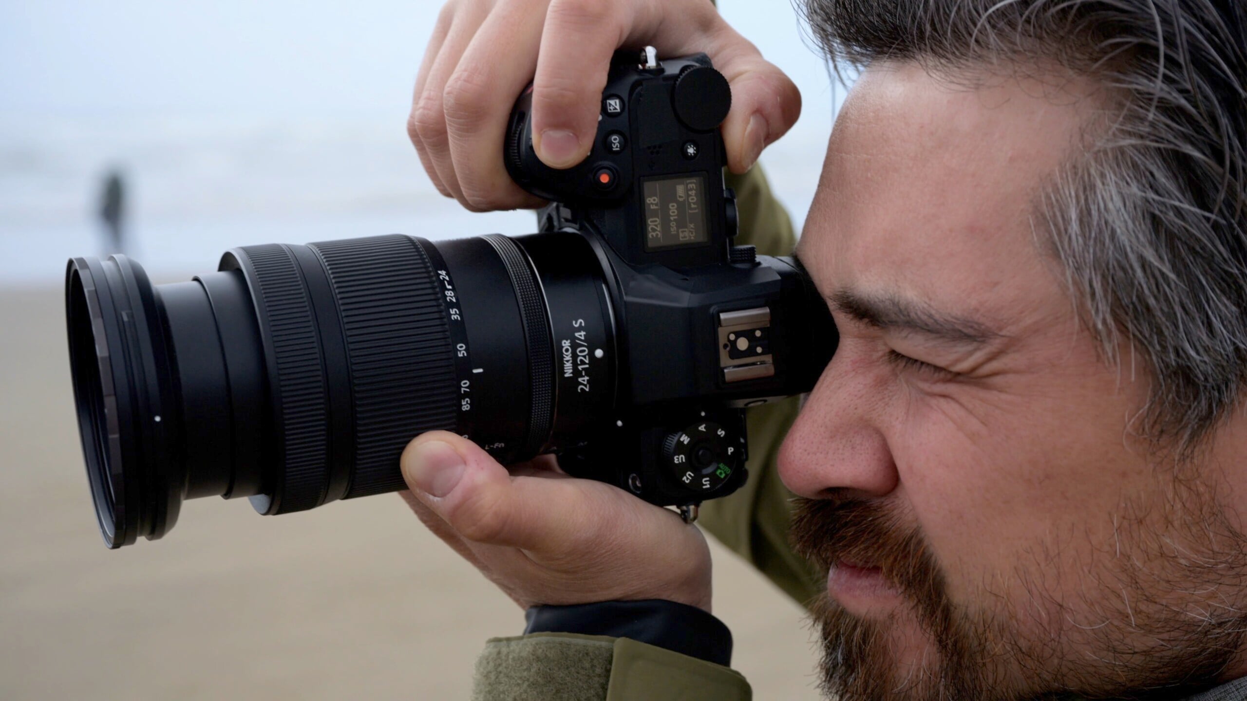 A man with a beard and short, graying hair focuses on taking a photograph with a digital camera fitted with a large lens. His left eye is closed, and he appears to be concentrating on capturing the perfect shot. The background is blurred and outdoors.