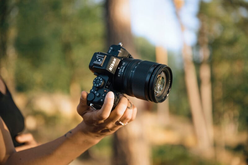 A person holds a Nikon DSLR camera outdoors with a blurred background of trees and sunlight.